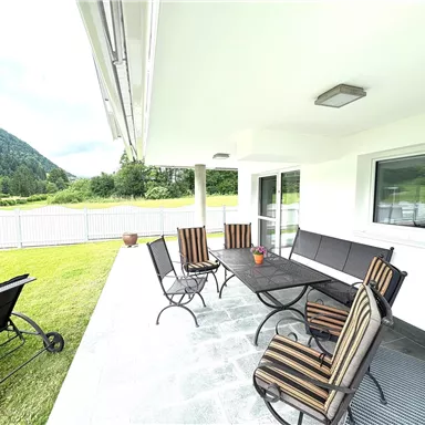 Ein gemütlicher Außenbereich mit einem Tisch und mehreren Stühlen. Im Hintergrund sind grüne Wiesen und Berge zu sehen.