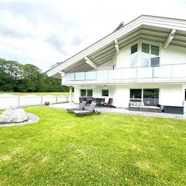 Ein modernes Haus mit großen Fenstern und einer geräumigen Terrasse. Der Garten ist gepflegt und bietet eine grüne Rasenfläche.