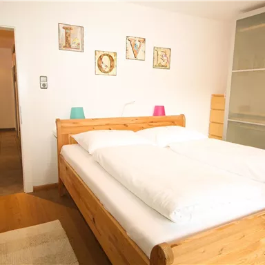 A cozy bedroom with a large wooden bed and white bedcovers. The walls are adorned with decorative pictures, and it has a bright, inviting character.