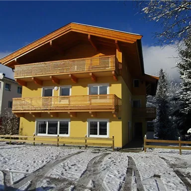 Ein gelbes Haus mit Holzbalkonen steht auf einer schneebedeckten Fläche. Im Hintergrund sind Bäume und blauer Himmel sichtbar.