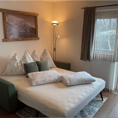 A cozy bedroom with a bed and several pillows. The room has large windows and a pleasant atmosphere.