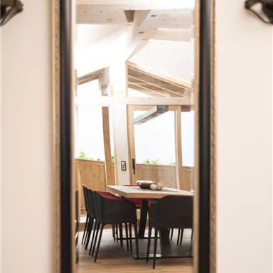 Ein großer Spiegel mit einem goldenen Rahmen spiegelt einen gemütlichen Essbereich wider. Es sind Stühle um einen Tisch angeordnet, die von Holz und Tageslicht umgeben sind.