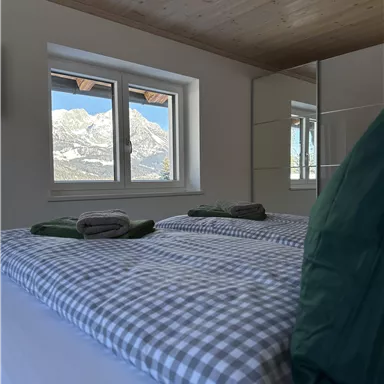 A cozy bedroom with a large window and a view of the mountains. The beds are decorated with checkered bedding and green cushions.