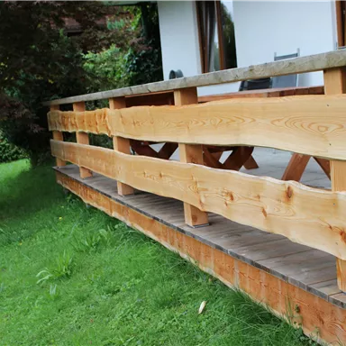 Ein Holzgeländer auf einer Terrasse umgeben von grünem Gras. Im Hintergrund ist ein Wohnhaus zu sehen.