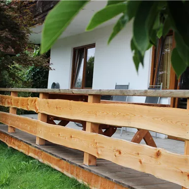 Eine Holzterrasse mit einem schönen Geländer umgibt das Haus. Im Hintergrund sind Fenster und grüne Pflanzen zu sehen.