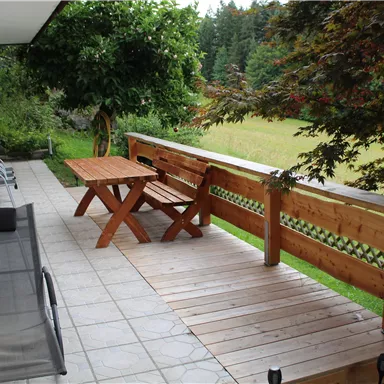Eine gemütliche Terrasse mit einem hölzernen Tisch und Stühlen. Umgeben von grünen Bäumen und einem weiten Feld.
