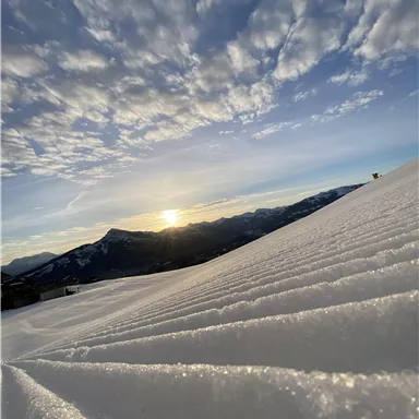 A snowy landscape with gentle hills illuminated by sunlight. The sky is clear and has some clouds.