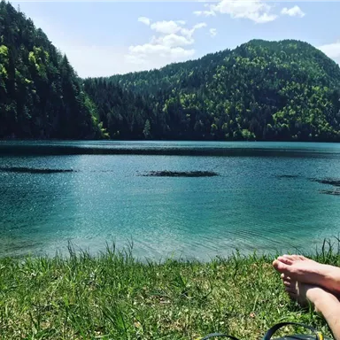 Ein ruhiger See umgeben von grünen Bergen und Bäumen. Im Vordergrund sind Füße auf dem Gras zu sehen.