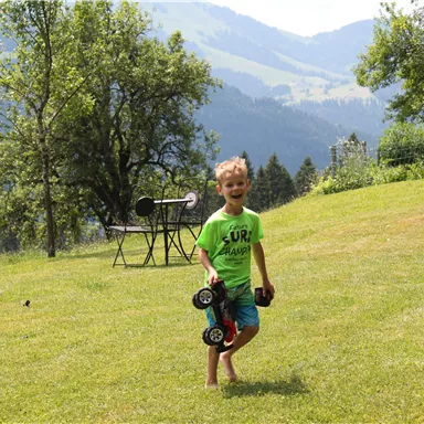 Ein Junge mit einem grünen T-Shirt spielt im Gras und hält ein Spielzeugauto in der Hand. Im Hintergrund sind Bäume und Berge zu sehen.