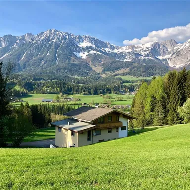 A picturesque landscape with snow-covered mountains in the background. In the foreground stands a beautiful house on a green meadow.
