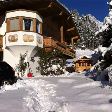 Ein charmantes Chalet im Schnee mit einem großen Balkon und umgeben von schneebedeckten Bäumen. Der Gartenweg ist deutlich sichtbar und der Himmel ist blau.