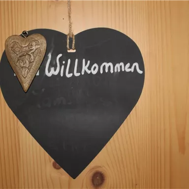 Ein herzförmiges Schild aus schwarzem Material mit der Aufschrift „Willkommen“. Es hängt an einer Holzoberfläche und hat einen kleinen Holzanhänger.