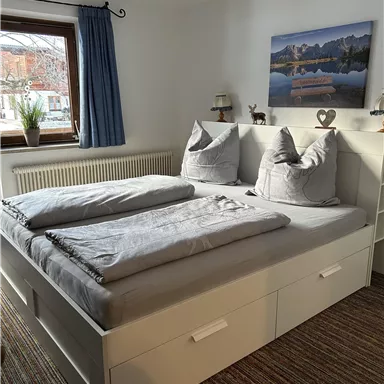 A cozy bedroom with a large double bed and two pillows. The room has a window with curtains and a wall decoration featuring a landscape picture.