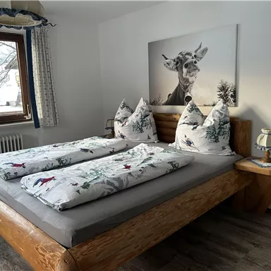 A cozy bedroom with a large wooden bed and decorative pillows. A picture of a deer hangs on the wall, and the window offers a view of the wintry landscape.