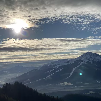 A breathtaking mountain landscape with snow-covered peaks and a wide valley below. The sun is shining through the clouds, creating a beautiful light atmosphere.