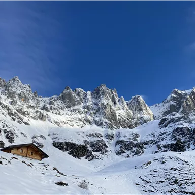 A snowy mountain landscape with impressive rocks and a clear blue sky. In the foreground stands a small cabin.