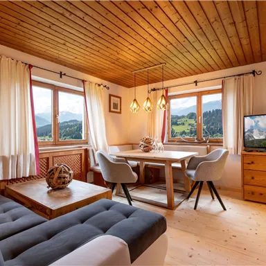 Ein gemütliches Wohn- und Esszimmer mit Blick auf die Berge. Helle Fenster, Holzdecke und moderne Möbel schaffen eine einladende Atmosphäre.