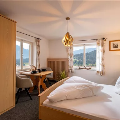 Ein gemütliches Zimmer mit einem Doppelbett und einem kleinen Tisch. Große Fenster bieten einen Blick auf die umliegenden Berge.