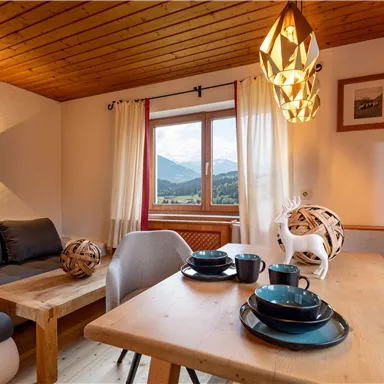 Ein gemütliches Wohnzimmer mit einem Holzstuhl und einem Tisch. Durch das Fenster ist eine wunderschöne Berglandschaft zu sehen.