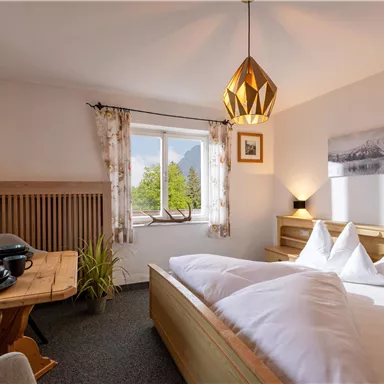 Ein helles Schlafzimmer mit einem Doppelbett und gemütlicher Einrichtung. Am Fenster sieht man einen Blick auf die Natur und eine Holztafel mit Geschirr.