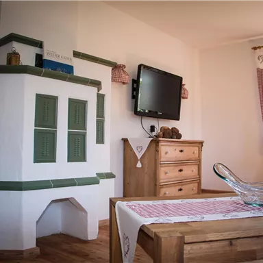 Ein gemütliches Wohnzimmer mit einem traditionellen Kachelofen und einem Fernseher. Der Holztisch ist mit einer Tischdecke und einer Glasschale dekoriert.