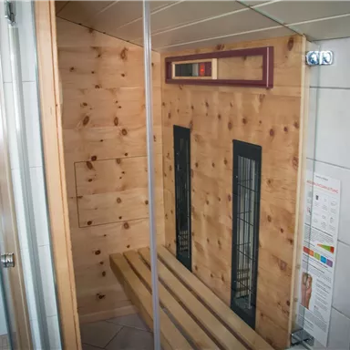 Eine moderne Sauna mit hellem Holzinterieur und zwei Heizpaneelen. Es gibt eine Sitzbank und geflieste Wände neben einer Duschkabine.