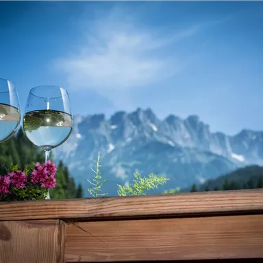 Zwei Gläser Weißwein stehen auf einem Holztisch mit Blick auf majestätische Berge. Im Vordergrund wachsen bunte Blumen.