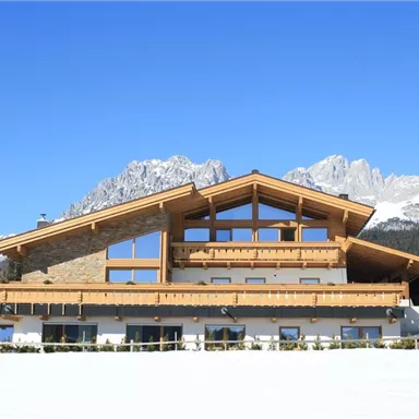 Ein modernes Holzhaus in den Bergen, umgeben von Schnee und majestätischen Gipfeln. Der klare blaue Himmel vervollständigt die idyllische Winterlandschaft.