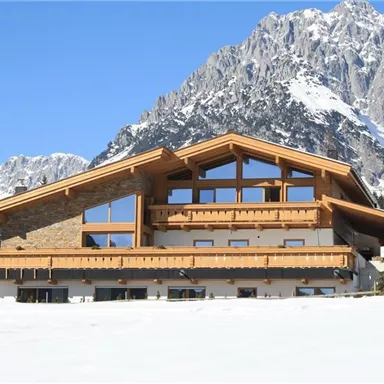 Ein modernes, alpines Chalet vor einer beeindruckenden Berglandschaft. Der schneebedeckte Boden und der klare Himmel vervollständigen die malerische Szenerie.