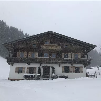 A traditional wooden house in the snow, surrounded by a wintry landscape. Gentle hills and trees are visible in the background.