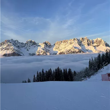 A picturesque winter landscape with snow-covered mountains and a dense layer of fog in the valley. The sky is blue and the sunrise illuminates the peaks.