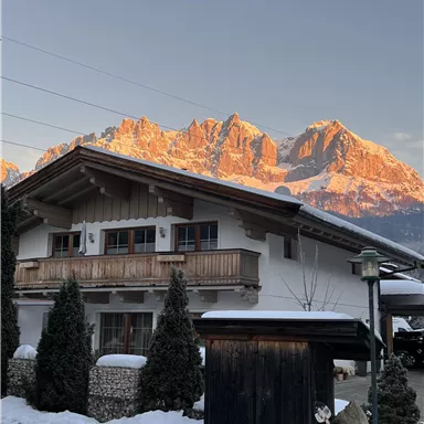 A beautiful house with a balcony and a snow-covered front yard. In the background, majestic mountains rise, illuminated by the evening sun.