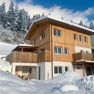 Ein modernes Haus aus Holz im Schnee, umgeben von schneebedeckten Bäumen. Die klare Winterlandschaft verleiht eine gemütliche Atmosphäre.