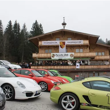 Eine Gruppe von Porsche-Fahrzeugen parkt vor einem Gebäude mit Holzfassade. Im Hintergrund steht eine Versammlung des Porsche Clubs.