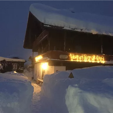 Ein winterliches Chalet bei Abenddämmerung, umgeben von hohem Schnee. Lichter verleihen dem Gebäude eine gemütliche Atmosphäre.