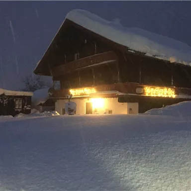 Ein rustikales Holzhaus im Schnee mit leuchtenden Lichtern. Die Umgebung ist von einer dicken Schneedecke bedeckt und der Himmel ist dunkel.