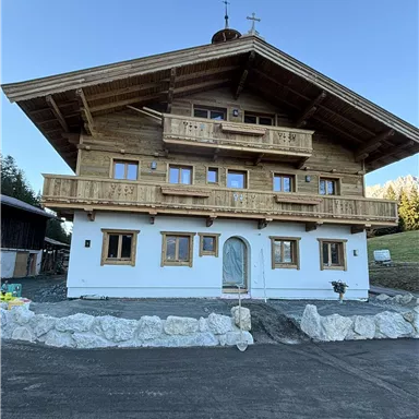 A traditional wooden house with a balcony in alpine style. The façade is partly painted white, and there is a well-kept outdoor area.