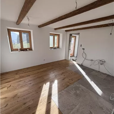 A bright room with wooden flooring and exposed wooden beams. The walls are still incomplete and there are windows that let in a lot of light.