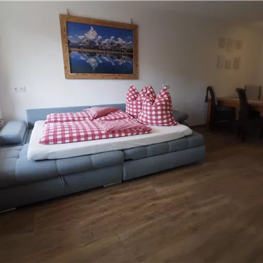 Ein gemütlicher Wohnraum mit einem großen Bett und bunten Kissen. Im Hintergrund hängt ein Landschaftsbild an der Wand.