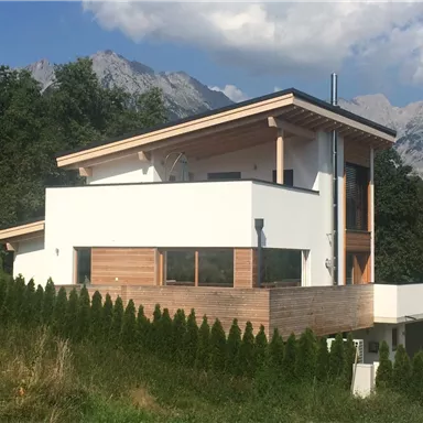 A modern house with wooden and plaster details stands in a green landscape. In the background are majestic mountains and a blue sky.