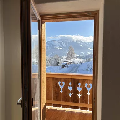An open window with a view of snow-covered mountains and a wooden balcony. The view is clear and sunny.