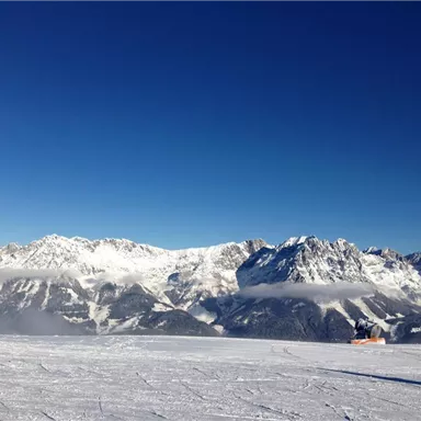Eine schneebedeckte Berglandschaft mit klar blauem Himmel. Ein Schneesportler fährt auf der Piste in die majestätische Bergkulisse.