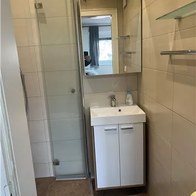 Ein modernes Badezimmer mit einer Dusche und einem Waschbecken. Die Wände sind hell gefliest und es gibt eine große Spiegelfläche.