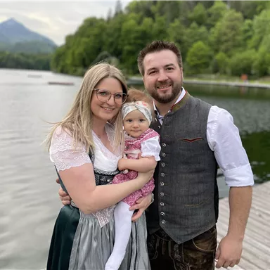 Eine Familie steht am Ufer eines ruhigen Sees, umgeben von grünen Bäumen. Sie tragen traditionelle bayerische Trachten und lächeln freundlich in die Kamera.