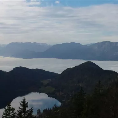Ein atemberaubender Bergblick mit einem glitzernden See und einer mystischen Nebelbank im Tal. Die umliegenden Berge sind leicht bewölkt und verleihen der Szenerie eine ruhige Atmosphäre.