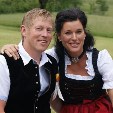 Ein Paar in traditioneller Tracht lächelt auf einer Wiese. Im Hintergrund sind Bäume und eine grüne Landschaft zu sehen.