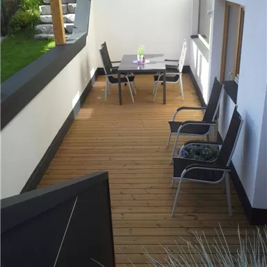 Eine einladende Terrasse mit Holzboden und einem Tisch sowie Stühlen. Die Umgebung ist grün und gepflegt.