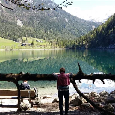 A quiet lake, surrounded by mountains and trees. One person stands on the shore while another sits on a bench enjoying the scenery.