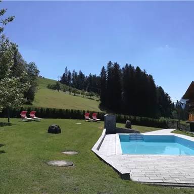 Ein schöner Garten mit einem Pool und Liegestühlen. Im Hintergrund sind grüne Hügel und Wälder zu sehen.