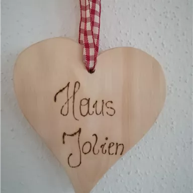 A heart-shaped wooden plaque with the inscription "Haus Jolien". It is equipped with a red checked ribbon for hanging.